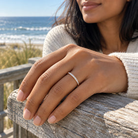 A yellow gold diamond ring sits on a hand resting on a weathered wooden railing with the ocean behind.