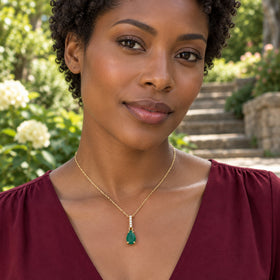 A woman in a burgundy blouse wears a yellow gold necklace with a green pear-shaped pendant beside garden steps.