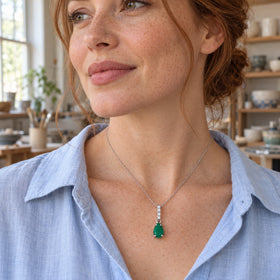 A freckled woman wears a silver necklace with a green pear-shaped pendant over a pale blue shirt in a ceramics studio.