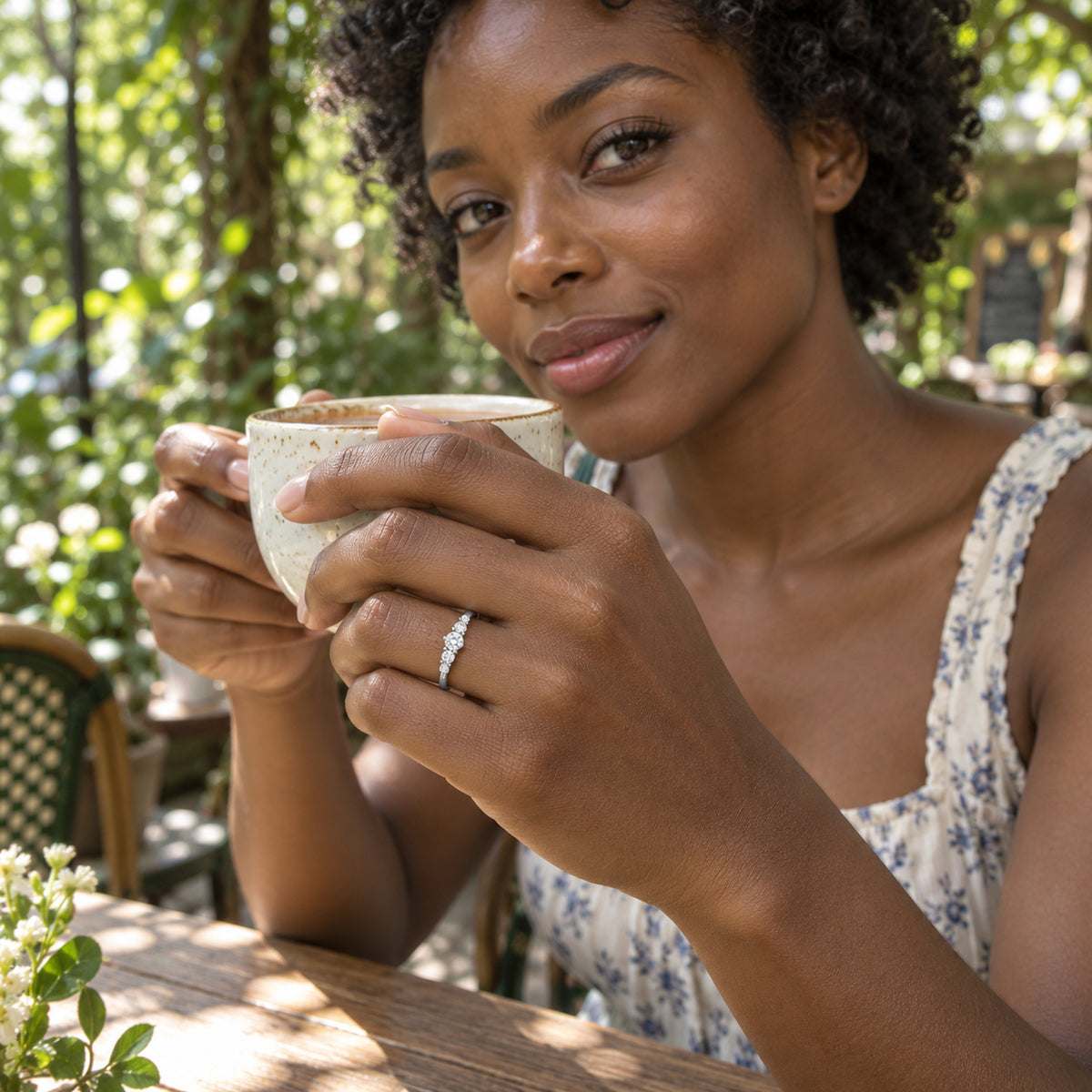 A woman wears a silver ring with five clear stones while holding a ceramic cup at a leafy outdoor table.