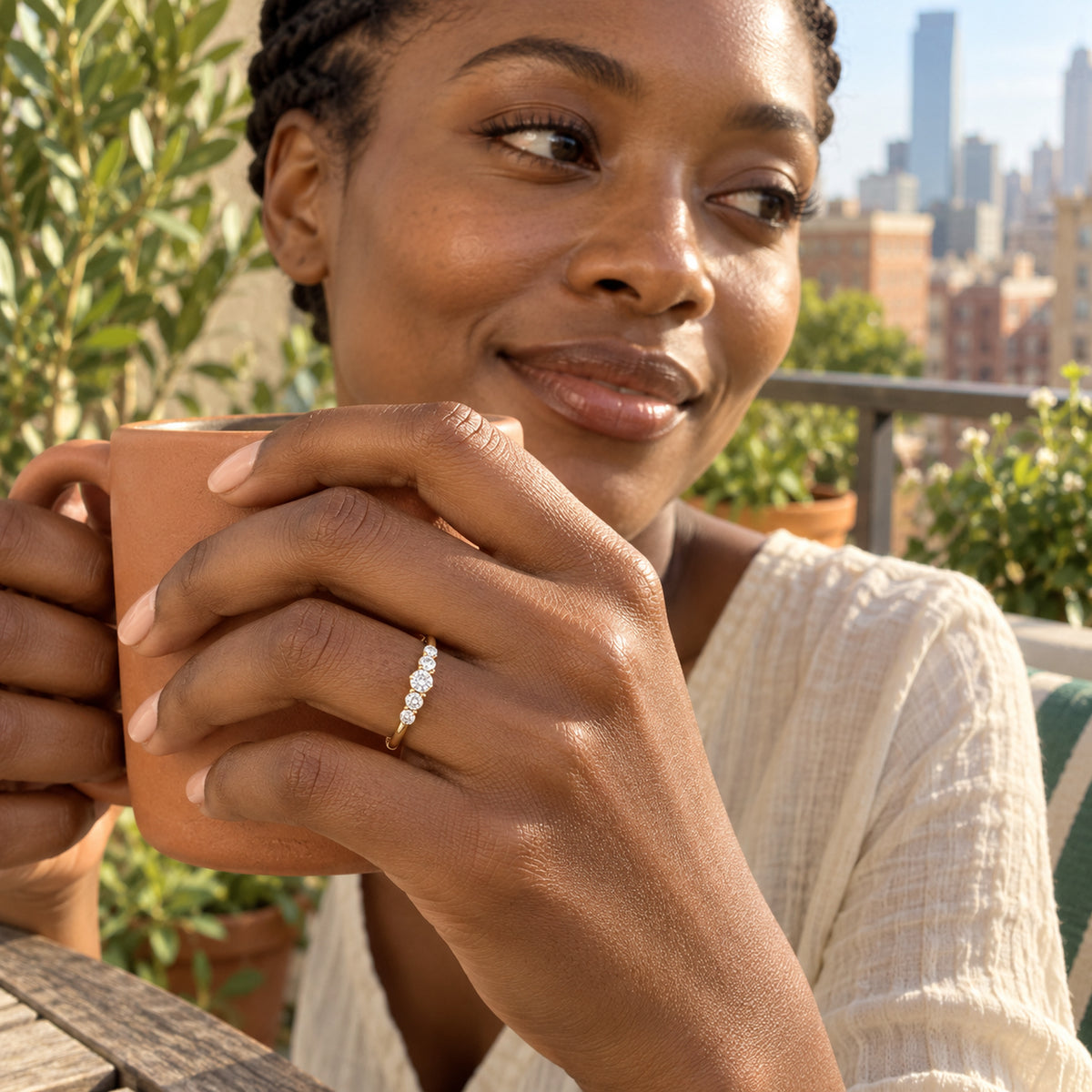 A yellow gold vermeil ring with five graduated white stones sits on a hand holding a terracotta mug on a planted balcony.