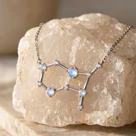 A silver constellation pendant with blue-white moonstones lies across a rough pale stone with its chain draped above.