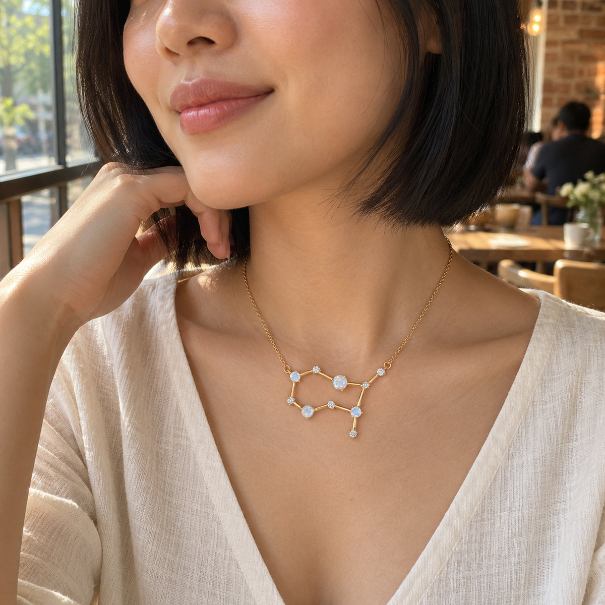 A woman with a black bob wears a yellow gold constellation necklace over a cream V-neck blouse beside a cafe window.