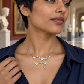 A woman with short dark hair wears a silver moonstone constellation necklace with a navy blouse in a museum corridor.