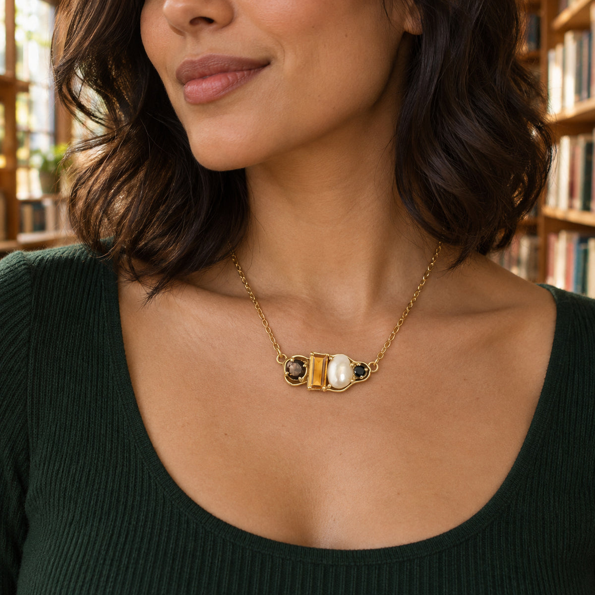 The yellow gold vermeil necklace rests at the collarbone of a woman in a green top with blurred bookshelves behind her.