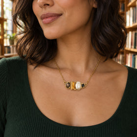 The yellow gold vermeil necklace rests at the collarbone of a woman in a green top with blurred bookshelves behind her.