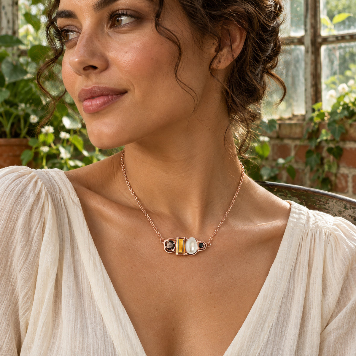 A woman in a cream blouse wears the rose gold vermeil citrine and pearl necklace beside leafy greenhouse windows.