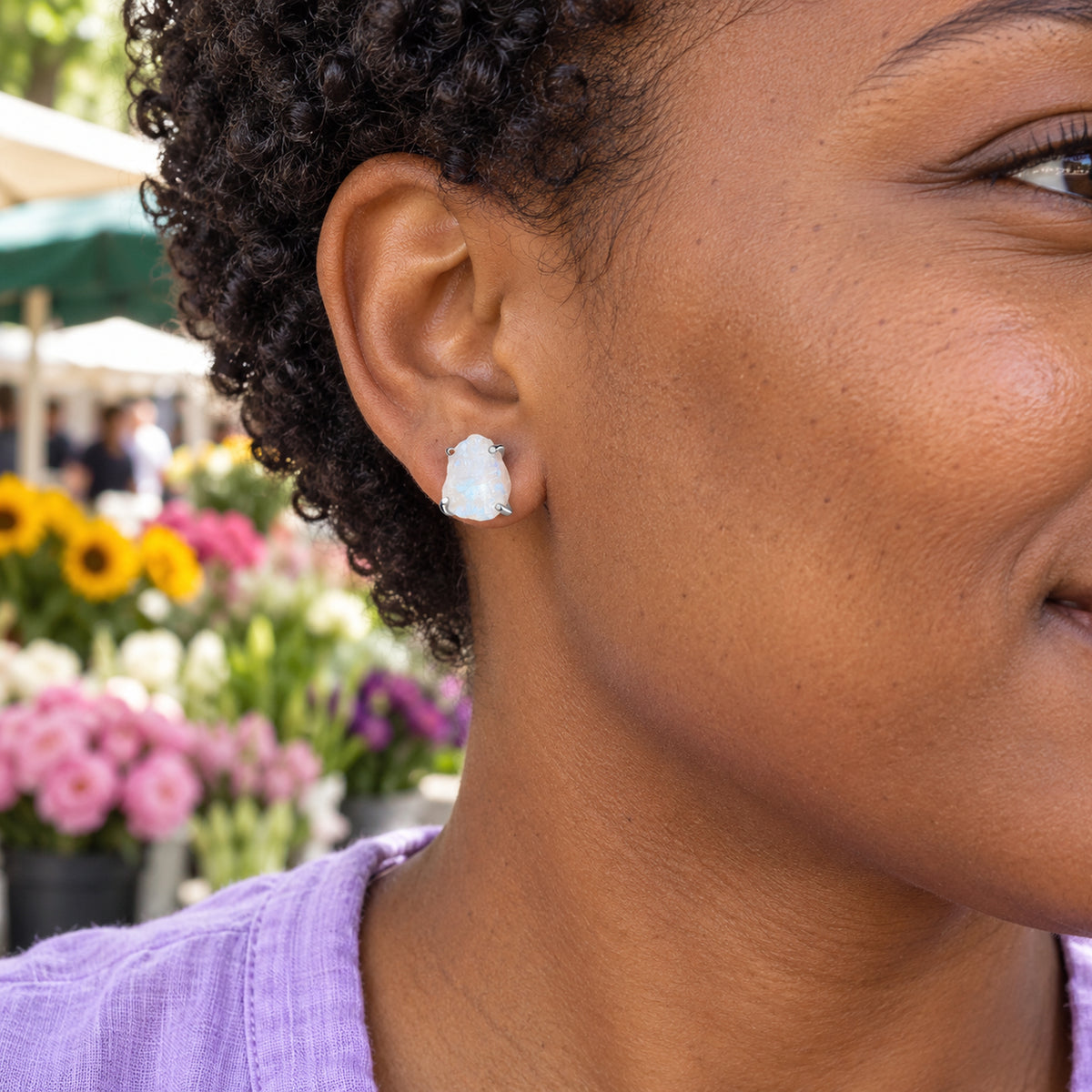 A silver moonstone stud is worn beside short curly hair at a colorful outdoor flower market.