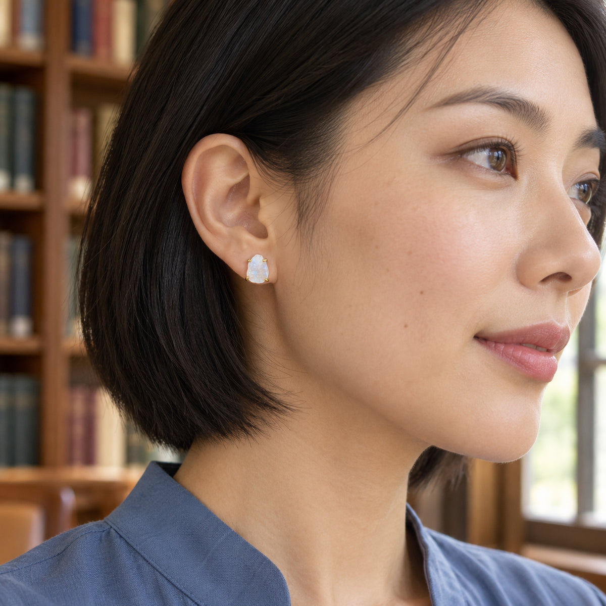 A yellow gold moonstone stud is visible below a black bob haircut with library shelves behind.