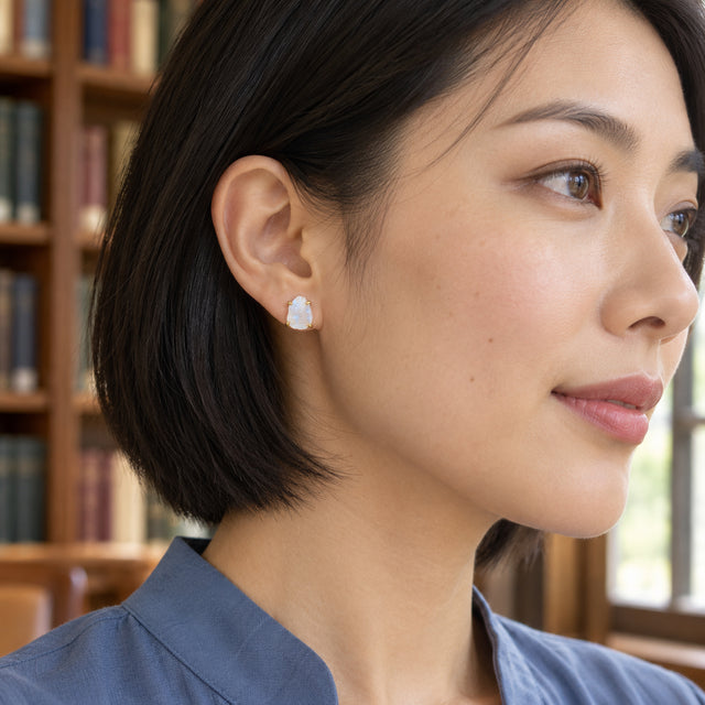 A yellow gold moonstone stud is visible below a black bob haircut with library shelves behind.