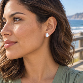 A rose gold moonstone stud is shown on an ear with chestnut hair and the sea in the background.