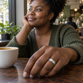 A woman in a green knit cardigan wears a sterling silver ring with white stones while resting her hand beside a cup on a wooden cafe table.