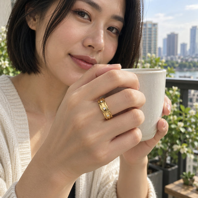 A yellow gold vermeil ring with a wavy band and three visible white stones is worn on a hand holding a ceramic mug on a balcony.