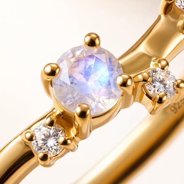 A pale faceted stone with blue reflections sits between rounded yellow gold prongs and clear accent gems.