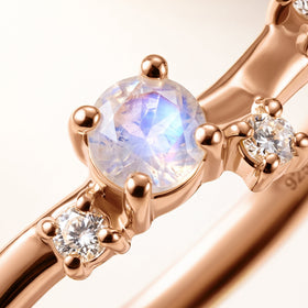 A blue-flashing round stone and two clear gems are shown closely in warm rose gold prong settings.