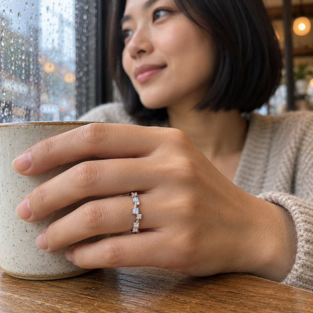 A silver ring with small pale stones is worn on a hand holding a ceramic cup beside a rain-speckled window.