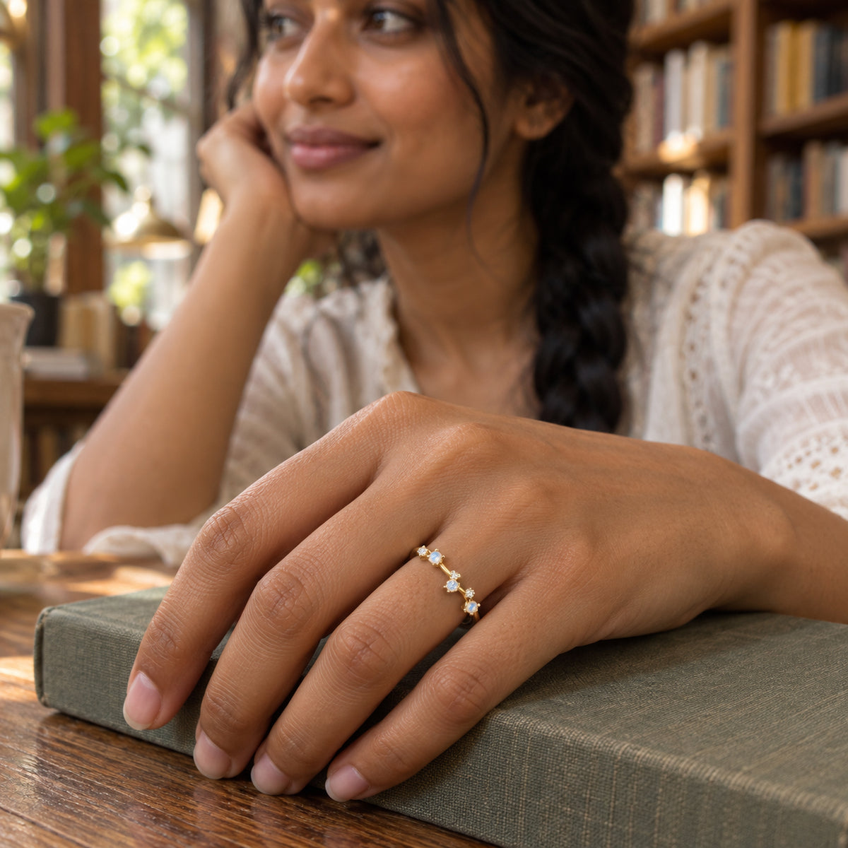 A yellow gold ring with blue-white and clear stones is worn on a hand resting on a cloth-covered book.