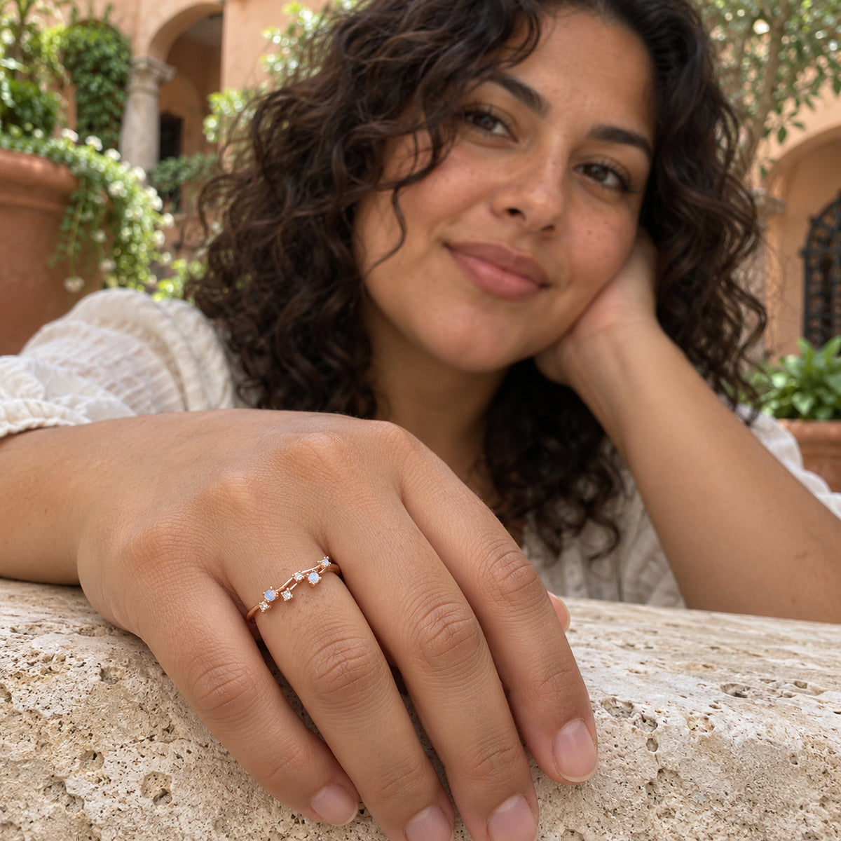A rose gold ring with small pale stones is worn on a hand resting on a textured stone ledge in a garden.