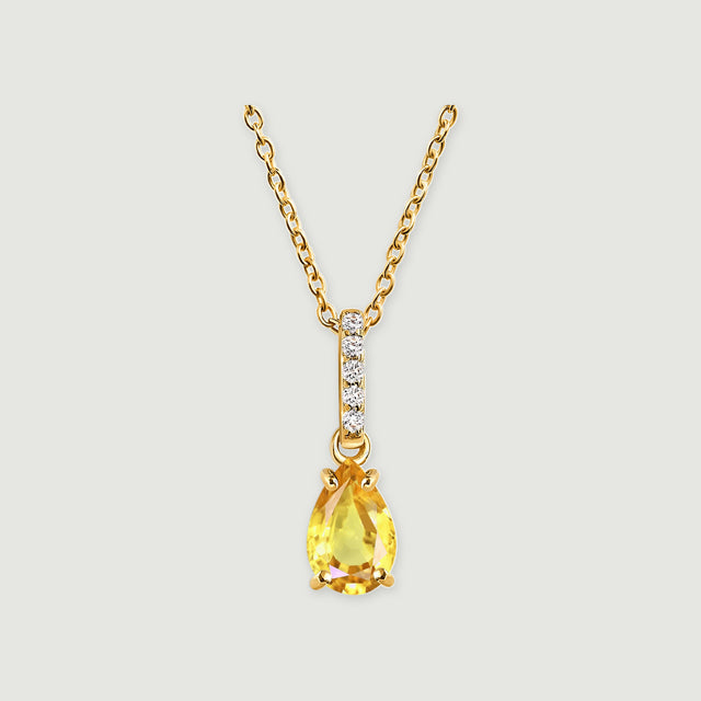 A pear-shaped yellow citrine hangs beneath a bar of five clear stones on a gold-toned chain against a light gray background.