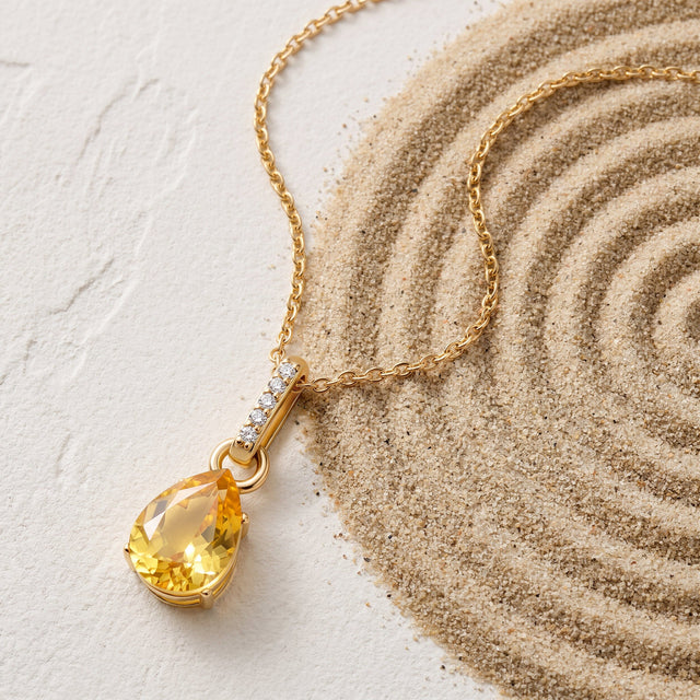 A gold-toned citrine necklace lies diagonally across a pale textured tabletop beside a sweep of fine sand.