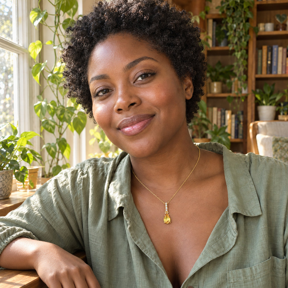 A woman in a sage shirt wears a gold-toned yellow citrine pendant beside a sunny window with plants and bookshelves.
