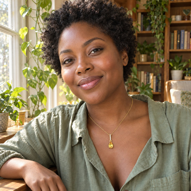 A woman in a sage shirt wears a gold-toned yellow citrine pendant beside a sunny window with plants and bookshelves.
