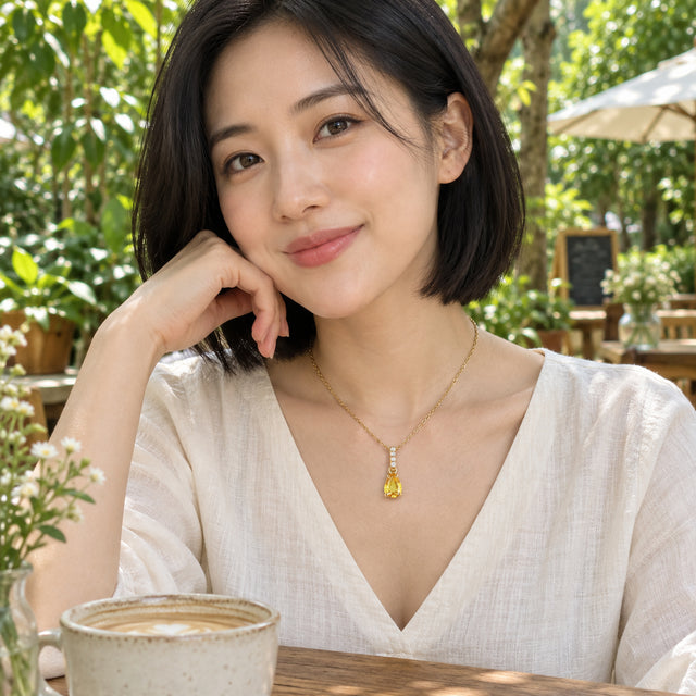 A woman with a black bob wears a yellow citrine necklace over a cream blouse at a leafy outdoor cafe.