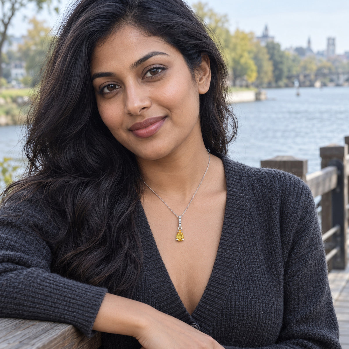 A woman in a charcoal cardigan wears a silver-toned yellow gemstone necklace beside a river and wooden railing.