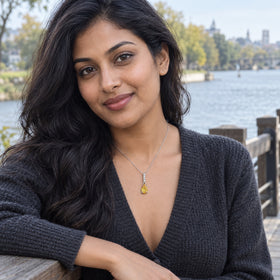 A woman in a charcoal cardigan wears a silver-toned yellow gemstone necklace beside a river and wooden railing.
