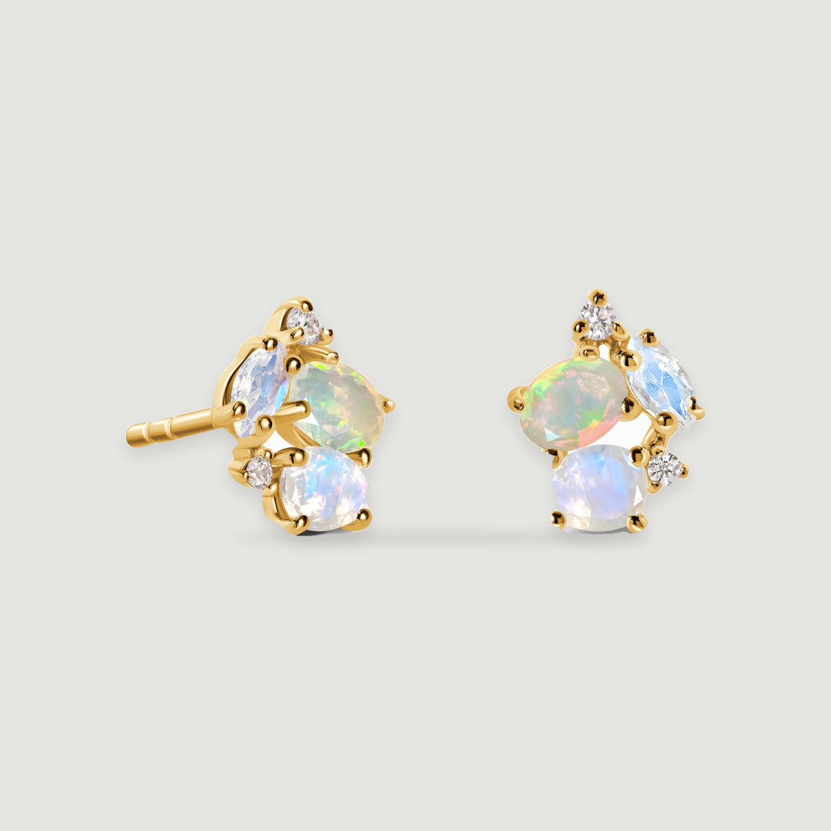 A pair of yellow gold stud earrings with iridescent oval and teardrop stones rests on a light gray surface.