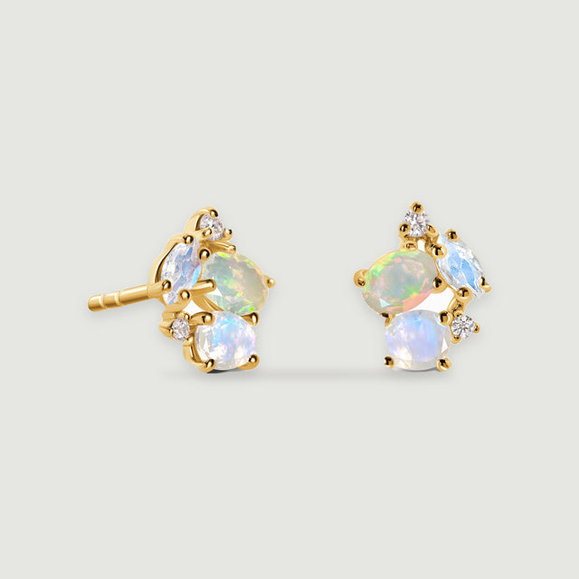 A pair of yellow gold stud earrings with iridescent oval and teardrop stones rests on a light gray surface.