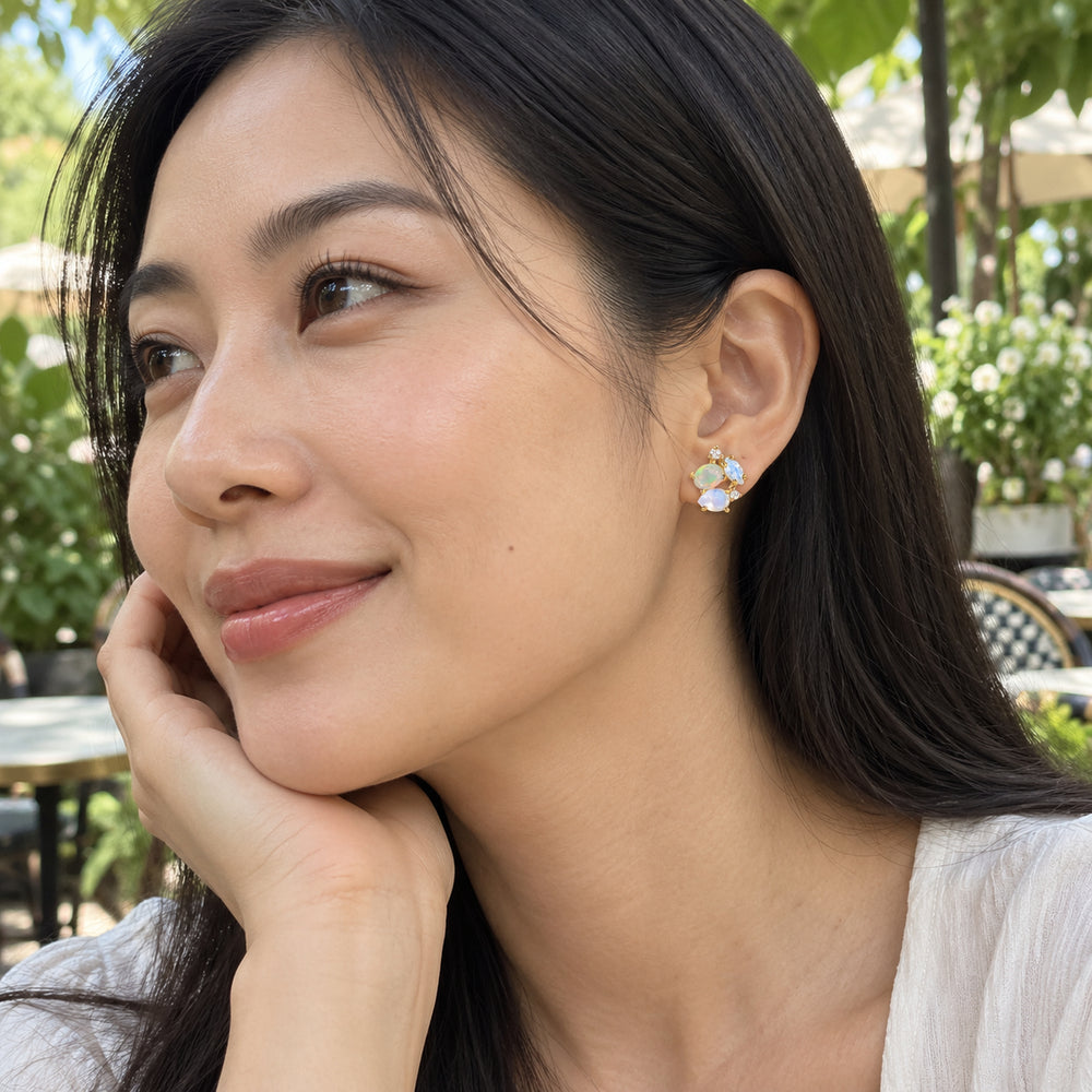 A woman with straight black hair wears a small yellow gold opal and pale gemstone stud at a leafy outdoor cafe.