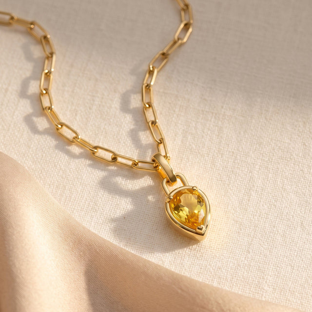 A gold plated citrine necklace rests diagonally on cream woven fabric beside a satin ribbon.