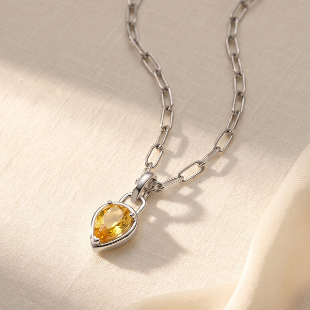 A white rhodium plated citrine pendant lies on cream fabric with its linked chain curving above it.