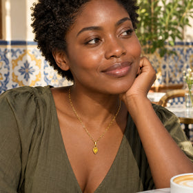 A woman with short curls wears a gold plated citrine necklace and an olive blouse in a tiled cafe.