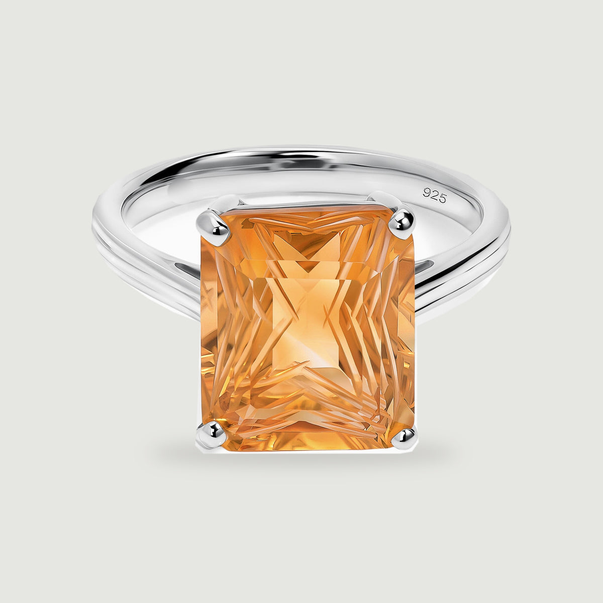 A rectangular golden citrine is held by four polished silver prongs above a slender ring band on a light gray surface.
