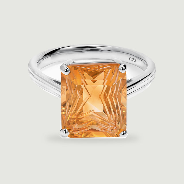 A rectangular golden citrine is held by four polished silver prongs above a slender ring band on a light gray surface.