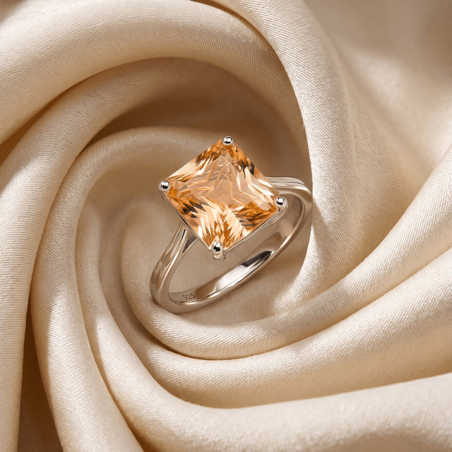 A silver citrine ring lies at an angle in the folds of ivory satin with its band and stone visible.