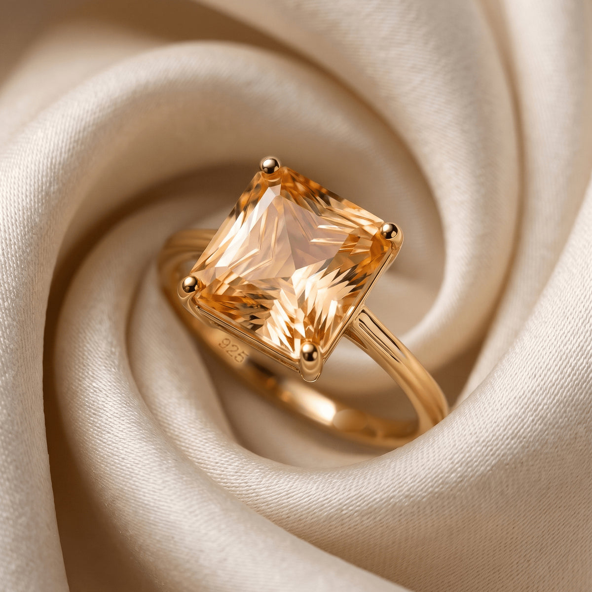 A yellow gold citrine ring rests among pale satin folds with four corner prongs framing its rectangular stone.