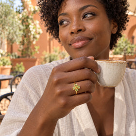 A yellow gold vermeil peridot clover ring is worn on a hand holding a cream ceramic cup in an outdoor cafe.