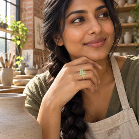 A woman with a dark braid wears a silver peridot clover ring on the hand resting beneath her chin in a pottery workspace.