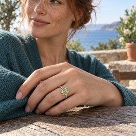 A silver ring with four green heart-shaped stones is worn on a freckled hand beside a teal cardigan and a coastal view.