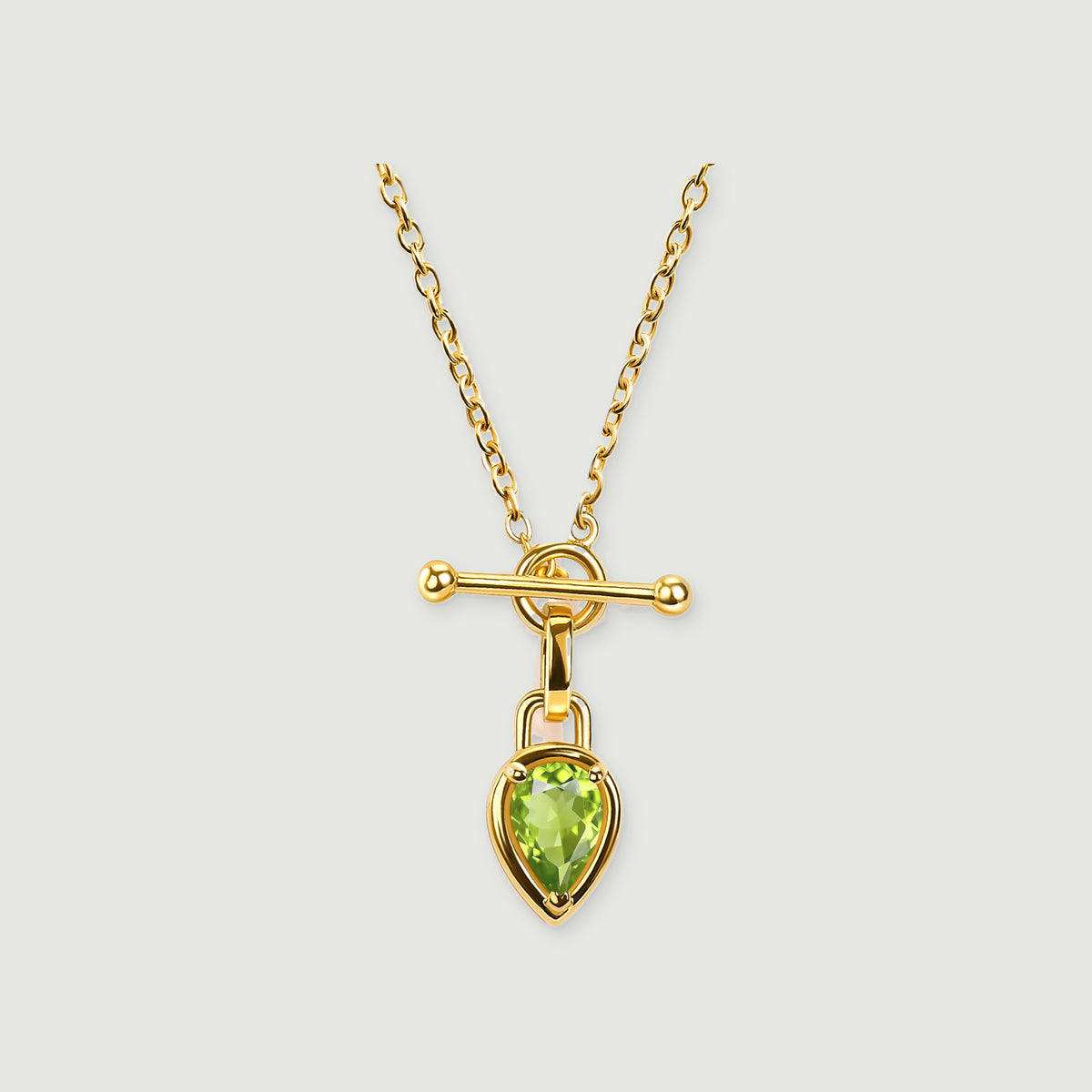 A yellow gold-tone necklace displays a green faceted pear pendant and a round-ended bar against pale grey.