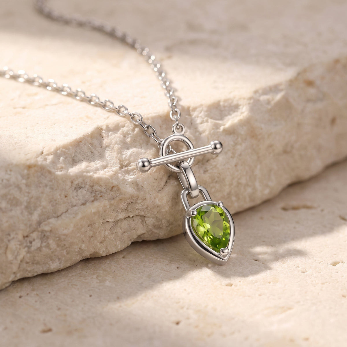 The silver-tone necklace lies across a cream stone ledge with its green pear pendant resting below the chain.