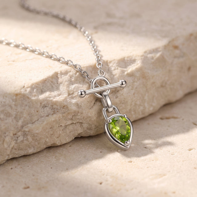 The silver-tone necklace lies across a cream stone ledge with its green pear pendant resting below the chain.