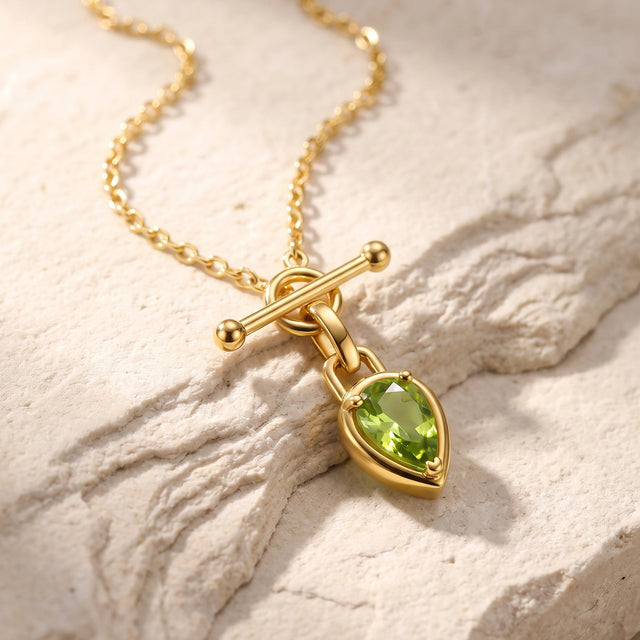 A gold-tone chain and green pear pendant rest diagonally on a textured cream stone surface.