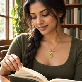 A woman in an olive top wears the gold-tone green pendant necklace while reading beside a sunny window.