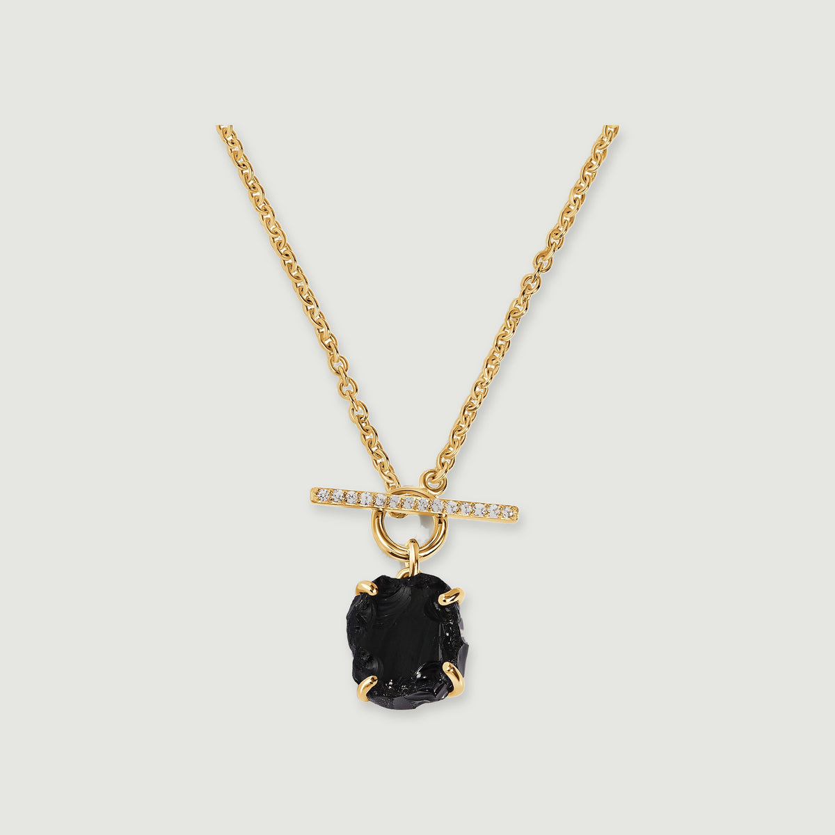 A yellow gold vermeil chain holds a rough black obsidian stone beneath a sparkling horizontal bar on a soft gray surface.