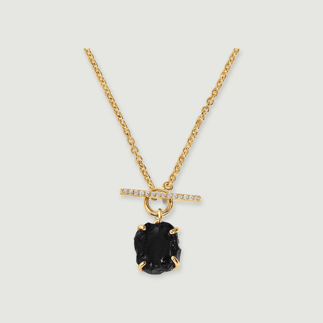 A yellow gold vermeil chain holds a rough black obsidian stone beneath a sparkling horizontal bar on a soft gray surface.