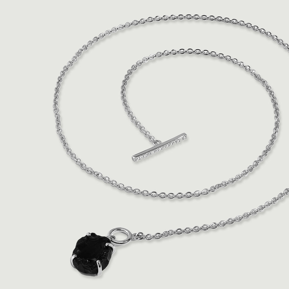 A silver necklace lies in loose curves with its crystal-set bar separated from the circular clasp beside a black obsidian pendant.